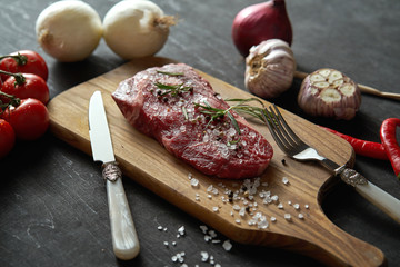 one raw piece of steak with salt, pepper on a wooden board and a set of vegetables on a black table
