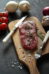 one raw piece of steak with salt, pepper on a wooden board and a set of vegetables on a black table