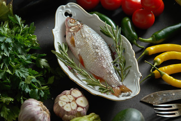 one fresh raw dorada fish in a white dish with a set of vegetables on a black table
