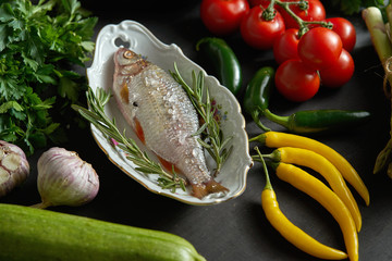 one fresh raw dorada fish in a white dish with a set of vegetables on a black table