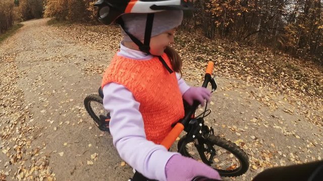 Take selfie on camera phone. One caucasian children rides bike road in autumn park. Girl riding black orange cycle in forest. Kid goes do bicycle sports. Biker motion ride with backpack and helmet.