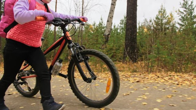 One caucasian children walk with bike in autumn park. Little girl walking black orange cycle in forest. Kid goes do bicycle sports. Biker motion ride with backpack and helmet. Mountain bike hardtail.