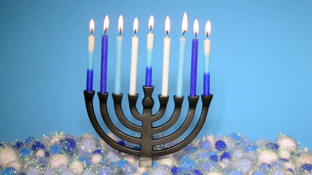 HD Video of a nine-branched menorah (also called a Chanukiah or Hanukiah) burning candles to celebrate Hanukkah against a light blue background with light and dark blue and white yarn balls underneath