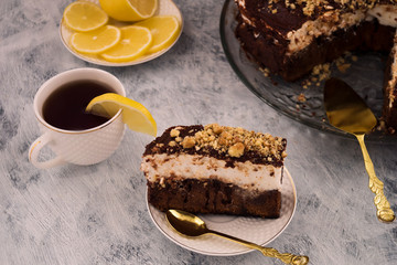 A piece of chocolate cake on a white plate. White and brown layer. A Cup of tea and a slice of lemon. Sliced lemon. Cutlery for dessert. Wooden background.