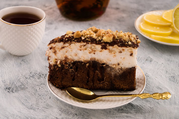 A piece of chocolate cake with grated nuts. A white layer of cream. Dishes for dessert. Tea in white mug. Light background.