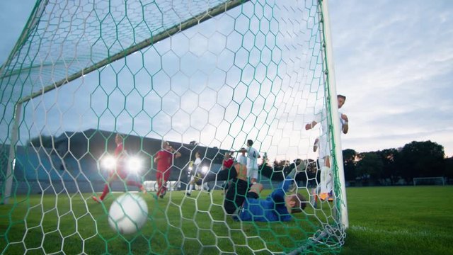 On Soccer Championship Player Kicks the Ball and Goalkeeper Tries to Defend Goals but Jumps and Fails to Catch the Ball. Camera Shot from Behind the Net with whole Stadium Visible.