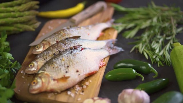 three fresh raw dorada fish on a wooden board with a set of vegetables on a black table