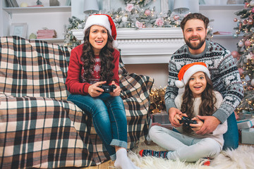 Small girl and young woman playing game by using joysticks. Father helps his daughter. They have...