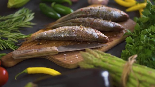 three fresh raw dorada fish on a wooden board with a set of vegetables on a black table
