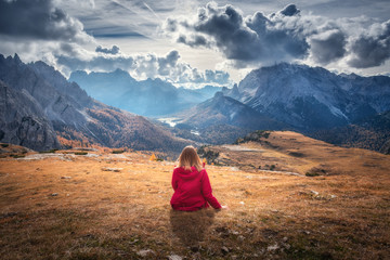Fototapeta premium Young woman is sitting on the hill against the majestic mountains at sunset in autumn in Dolomites, Italy. Landscape with girl, cloudy sky, orange grass, high rocks with forest in fall. Italian alps
