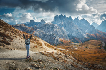 Young woman with raised up arms and majestic mountains at sunset in autumn in Dolomites, Italy. Landscape with happy girl, dramatic sky with clouds, orange trees, high rocks in italian alps in fall