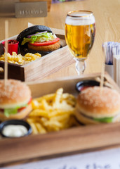 One black colored and two normal small mouth-watering, delicious burgers on the wooden tray served with red ketchup sauce and beer.