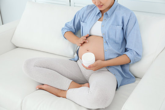 Pregnant woman applying body cream on belly at home, closeup
