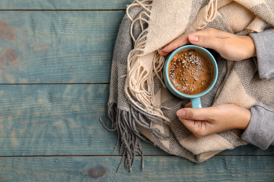 Woman with warm scarf holding cup of hot winter drink on wooden background, top view. Space for text