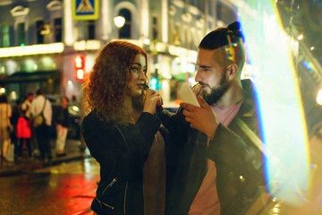 Couple in love on date. Boyfriend holding phone. She hugs him and smokes an electronic cigarette. They look at each other in street of night city. Wet asphalt after rain.