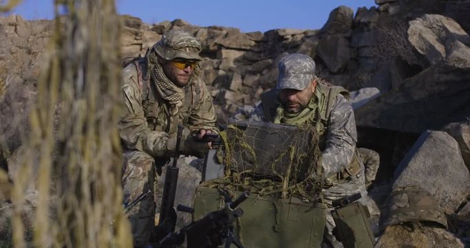Medium shot of fully equipped and armed soldiers looking at a computer and another looking at a handheld device in the field