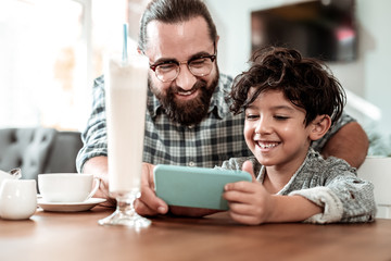 Game on phone. Cute handsome curly dark-haired son feeling entertained playing game on phone