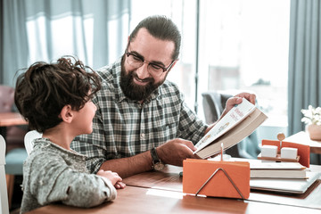 Lovely son. Happy dad wearing squared shirt looking at his lovely cute son reading book together