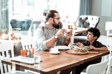 Lovely son. Beaming bearded man wearing glasses looking at his lovely son while eating pizza for lunch