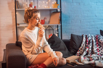 beautiful young blonde woman sitting on couch and eating gingerbread cookie at christmas time