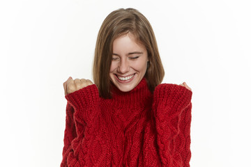 Success, joy and happiness concept. Happy excited young female in stylish warm sweater posing isolated keeping fists clenched and smiling broadly, rejoicing at good news, making her dream come true