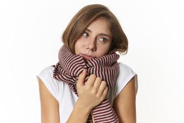 Obraz premium Picture of beautiful woman in white t-shirt warming up being wrapped in woolen striped scarf, posing against blank studio wall background with copyspace for your text or advertising content