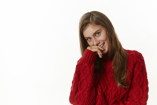 Flirtatious playful young Caucasian woman wearing stylish knitted long sleeved sweater smiling joyfully, holding hand on her lips, posing at white blank studio wall with copyspace for your content