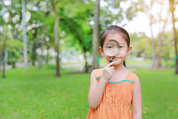 Fototapeta premium Pretty little Asian child girl with magnifying glass looks at flower in summer park.