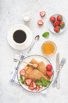 Crepes or blini with honey, strawberries and cup of coffee on white concrete background. Top view of tasty healthy breakfast