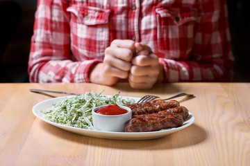 plate with grilled sausages and cabbage salad. Woman holds herself and her hands at a table in a restaurant in a red plaid shirt. copy space for design.