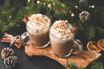 Zwei Tassen heiße Schokolade mit Schlagsahne und Zimt auf einem hölzernen Servierbrett, umgeben von Tannenbaum, Weihnachtsbeleuchtung, Zimt und Tannenzapfen. Gemütliches Weihnachtsgetränk