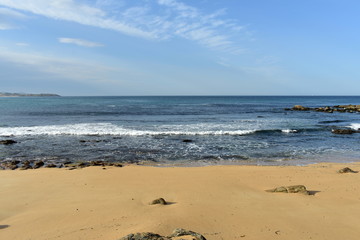 Beach with golden sand, rocks and blue water with waves and foam. Galicia, Spain, blue sky with clouds.