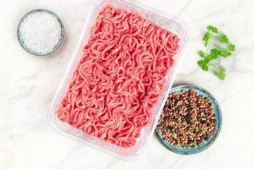 Minced meat in a plastic tray, shot from the top on a white marble background with salt, pepper,...
