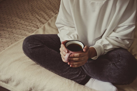Girl in a white sweater and gray jeans sitting on the bed with a cup of tea