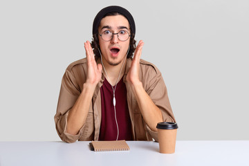 Shocked hipster guy keeps hands near cheeks, opens mouth widely, dressed in fashionable clothes, sits at white desk with organizer, listens news online with earphones, drinks aromatic beverage