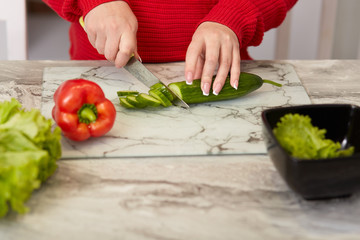 Cropped image of womans hands cuts vegetables with knife, slices cucumber, pepper and lettuce at kitchen table, prepares vegeterian fresh salad, dressed in red clothes, has manicure. Cooking concept