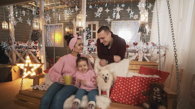 Mother, father, little daughter and white dog have fun sitting at Christmas bench. Young family in pullovers celebrating winter holidays at porch, expressing love and care in slow motion