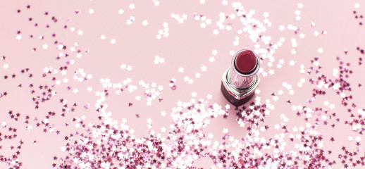 Red lipstick and holographic glitter confetti in the form of stars on pink background Flat lay top view copy space. Makeup accessories, holiday winter decoration
