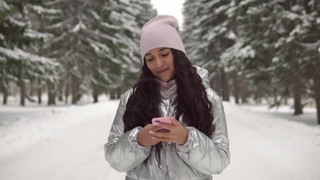 Attractive girl in a good mood walks through the winter forest in the afternoon and uses a smartphone. 4K
