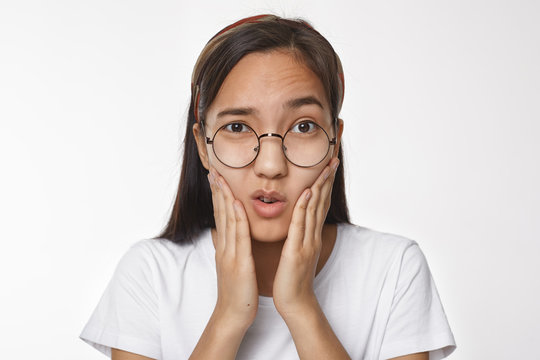 Studio shot of frustrated young Chinese female in round spectacles touching cheeks, having painful facial expression because of toothache. Nerdy Korean girl frowning, holding hands on face