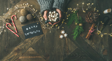 Christmas, New Year background. Flat-lay of greeting card, glittering toys, woman's hands holding mug of hot chocolate, cinnamon, scissors, light garland over wooden table, top view, copy space