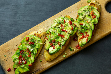 Three bruschetta with avocado slices, green onions, spices and chili peppers on a wooden tray against a black background.