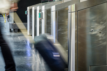 Side view of a man with a rolling carry-on luggage passing through stainless steel ticket gates in a metro station with motion blur.