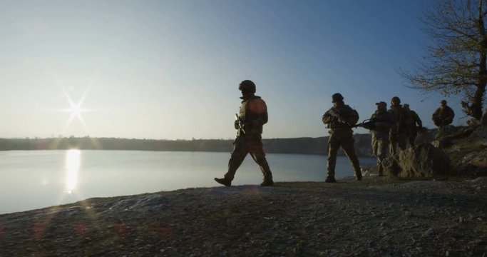 Medium slow motion shot of fully equipped and armed soldiers walking in single file by the side of a lake at sunset