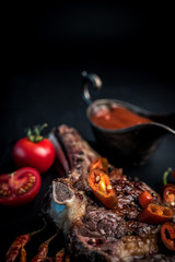 Fototapeta na wymiar Grilled hot ribeye beef steak on bone with spicy basting sauce and tomatoes on dark cooking background