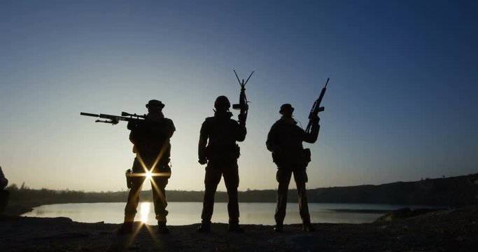 Medium shot of three soldiers posing with guns against the backdrop of a lake as the sun sets behind them