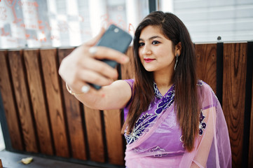 Indian hindu girl at traditional violet saree sitting at cafe table with mobile phone at hands,...