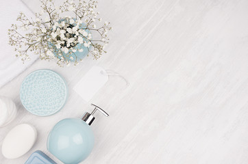 Elegant cosmetics set of accessories for beauty care - soap, towel, soap dispenser and circle pastel blue bowls, silver cosmetic bag, white flowers on white wood background, top view.