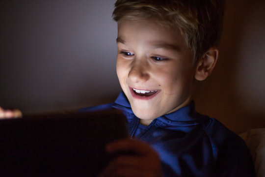 Cute kid with tablet in the dark. Child using tablet at night. Bright screen light reflex on kid face.