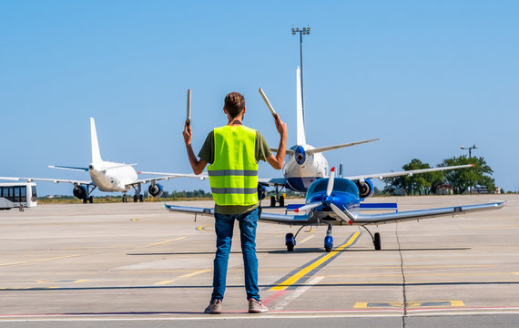 Dispatcher Geaturing Signs Directing The Blue Shiny Sport Plane On The Airport Runway